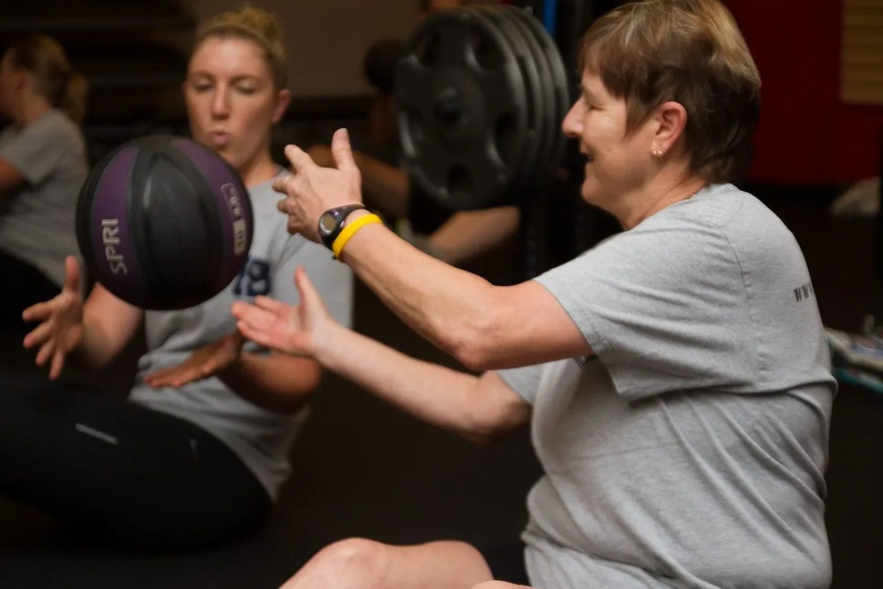 A coach and an older member trading medicine ball passes in the studio
