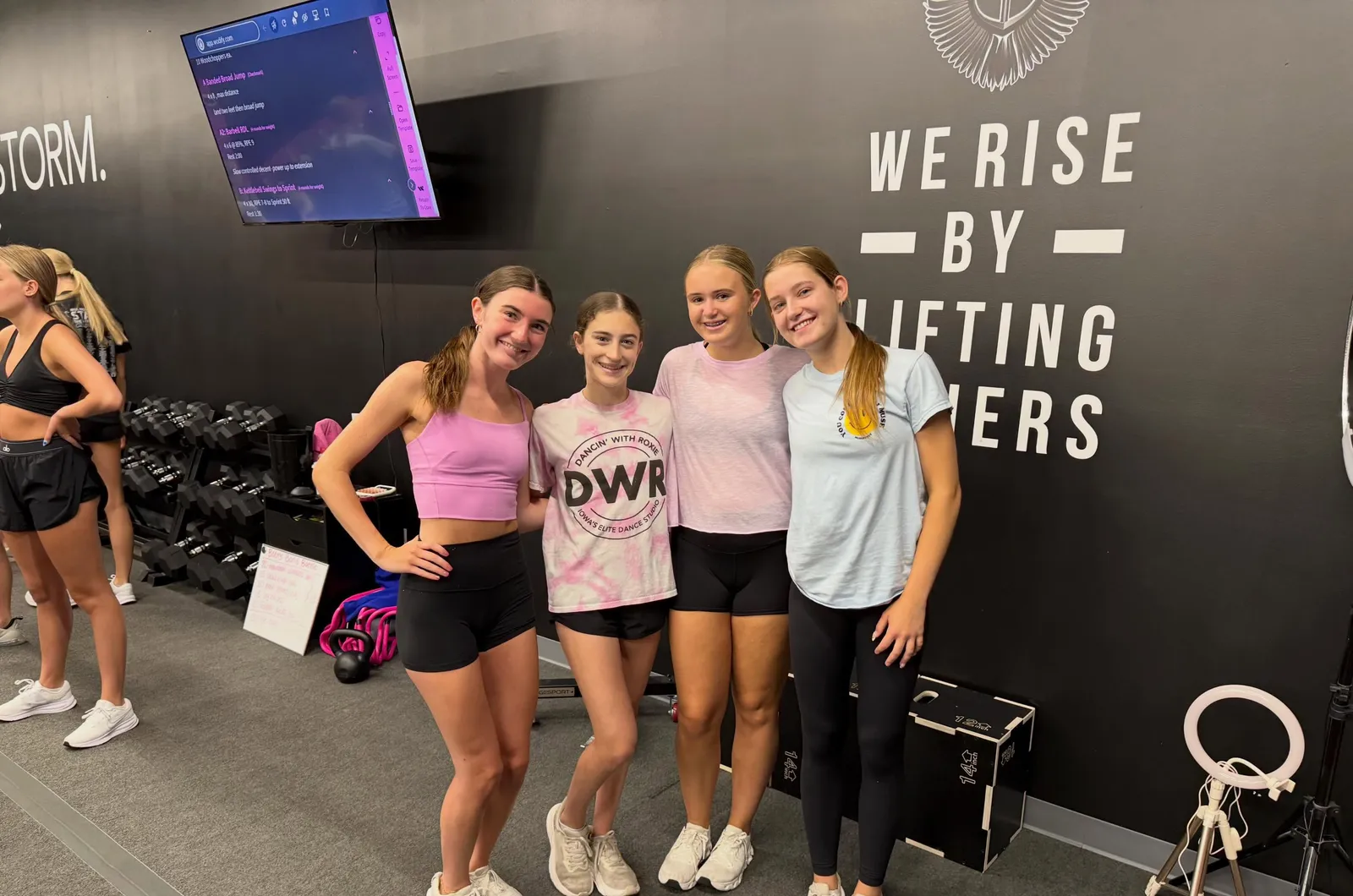 Four members smiling in front of the We Rise By Lifting Others wall at Valkyrie Society