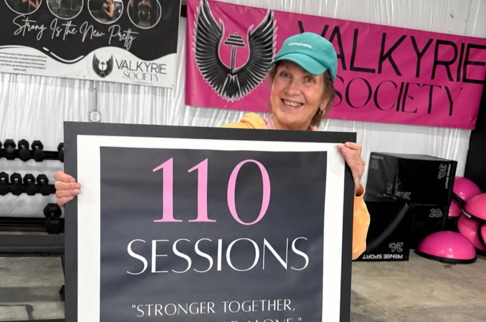 A smiling member holding her 110 sessions milestone sign under the Valkyrie Society banner