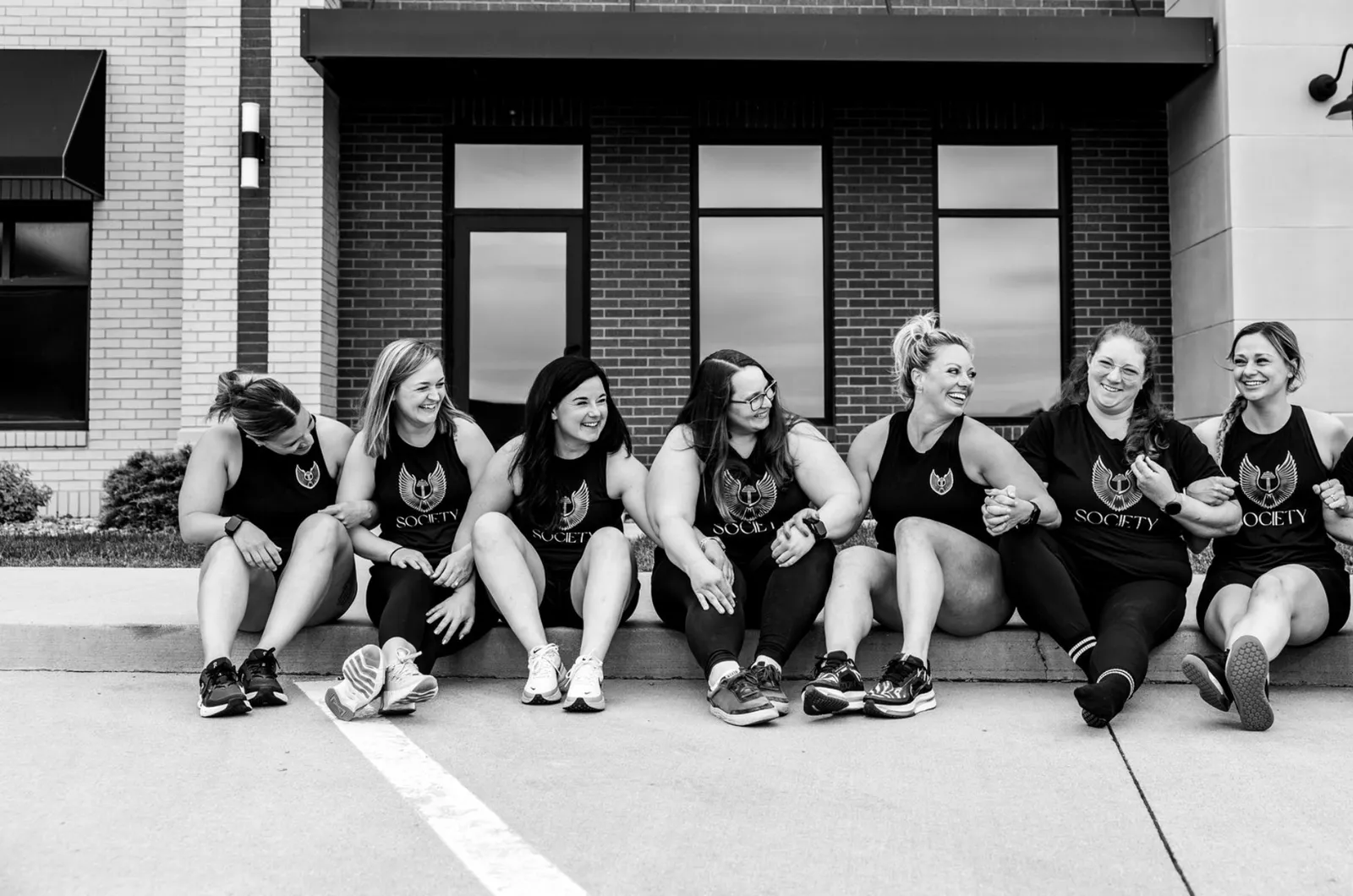 Seven members sitting together on the curb outside the gym, in black and white
